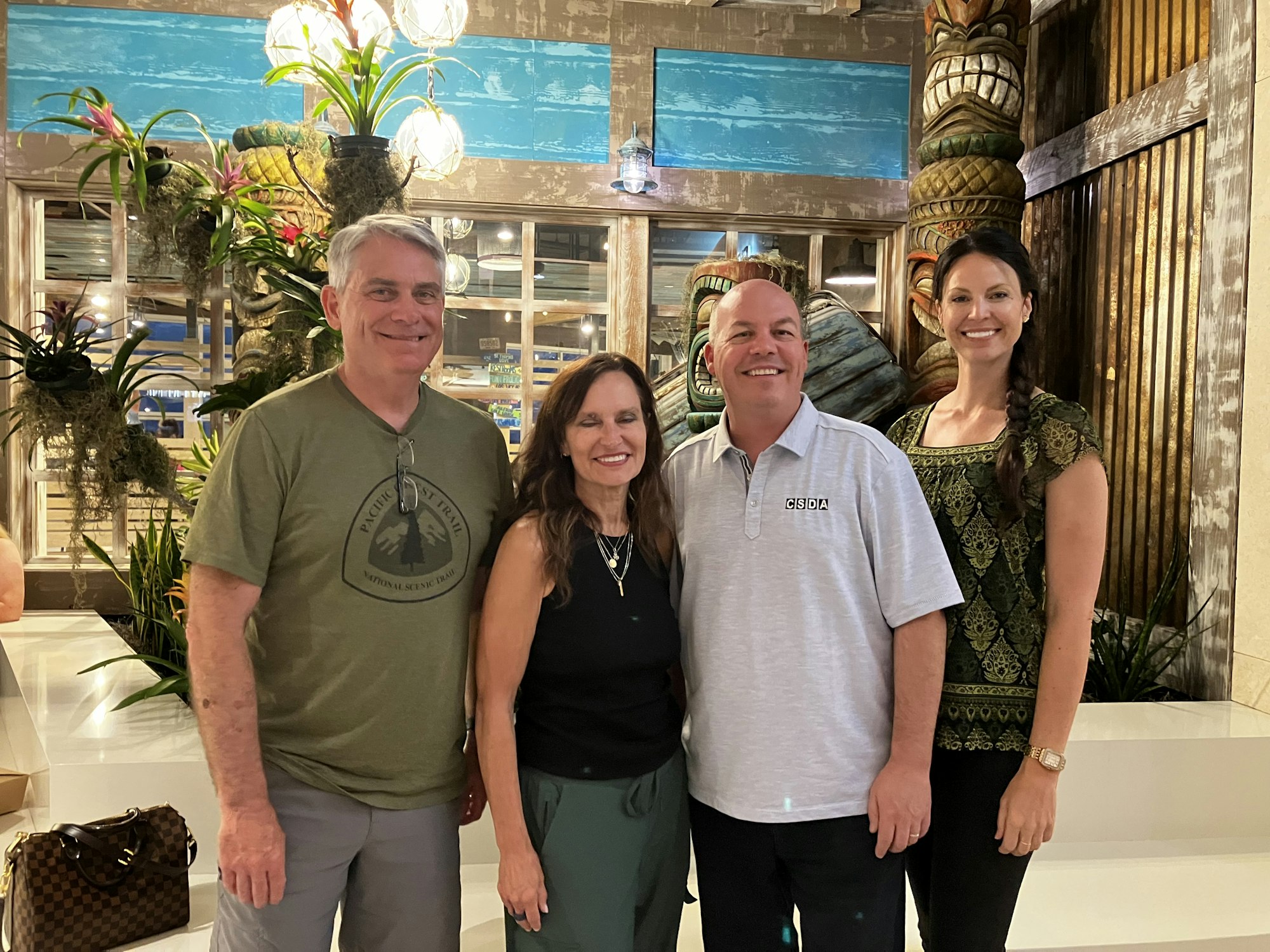 Four people smiling in a decorated indoor space with plants and tiki statues.