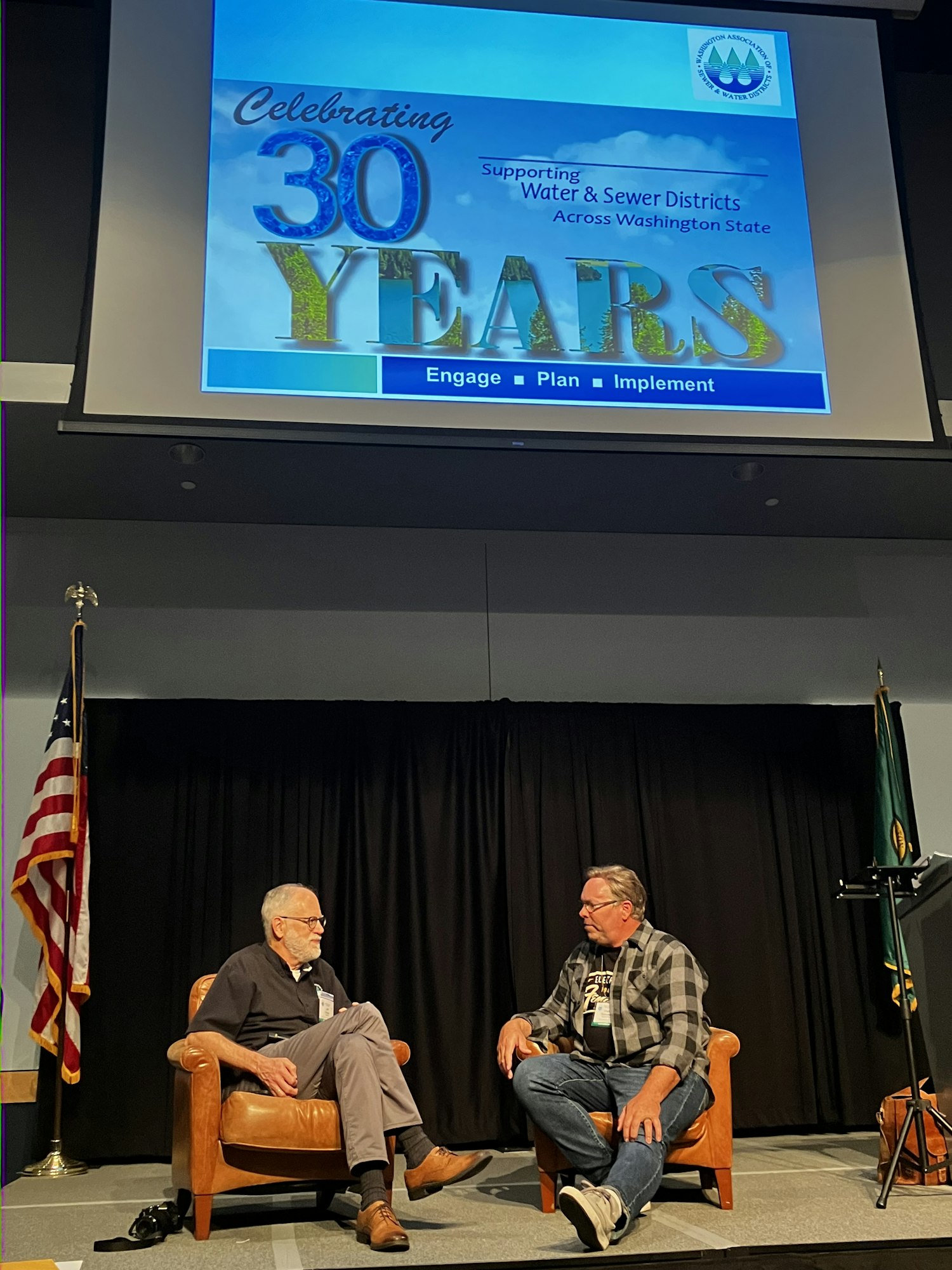 Two men seated on stage discussing, with a screen displaying "Celebrating 30 Years" related to water & sewer districts in Washington.