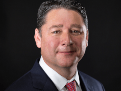 Tom Tight in a suit and red tie against a dark background.