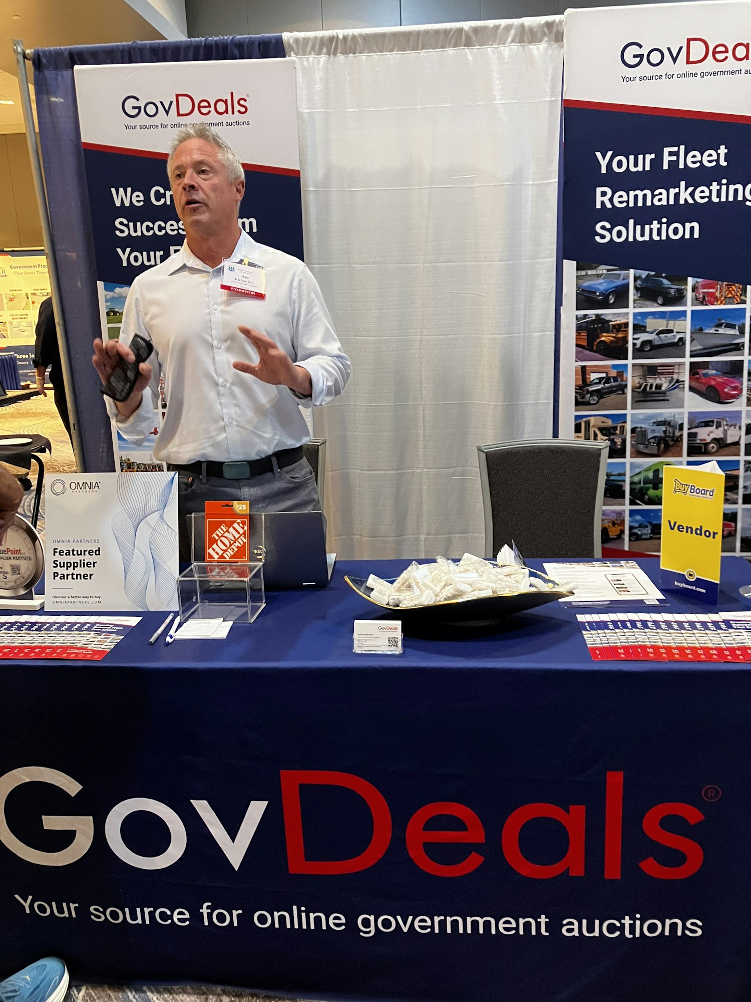 A man is at a GovDeals booth with promotional materials and a "Vendor" sign.