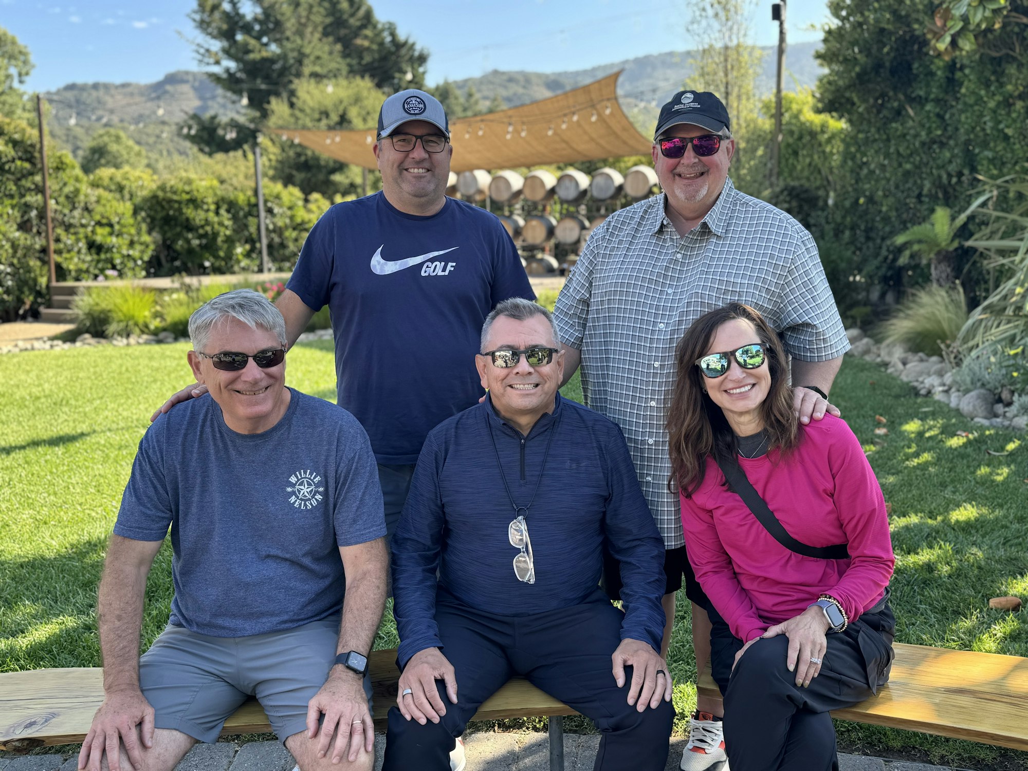 Five people in a park, sitting and standing on a bench, wearing casual clothes and sunglasses, with trees and barrels in the background.