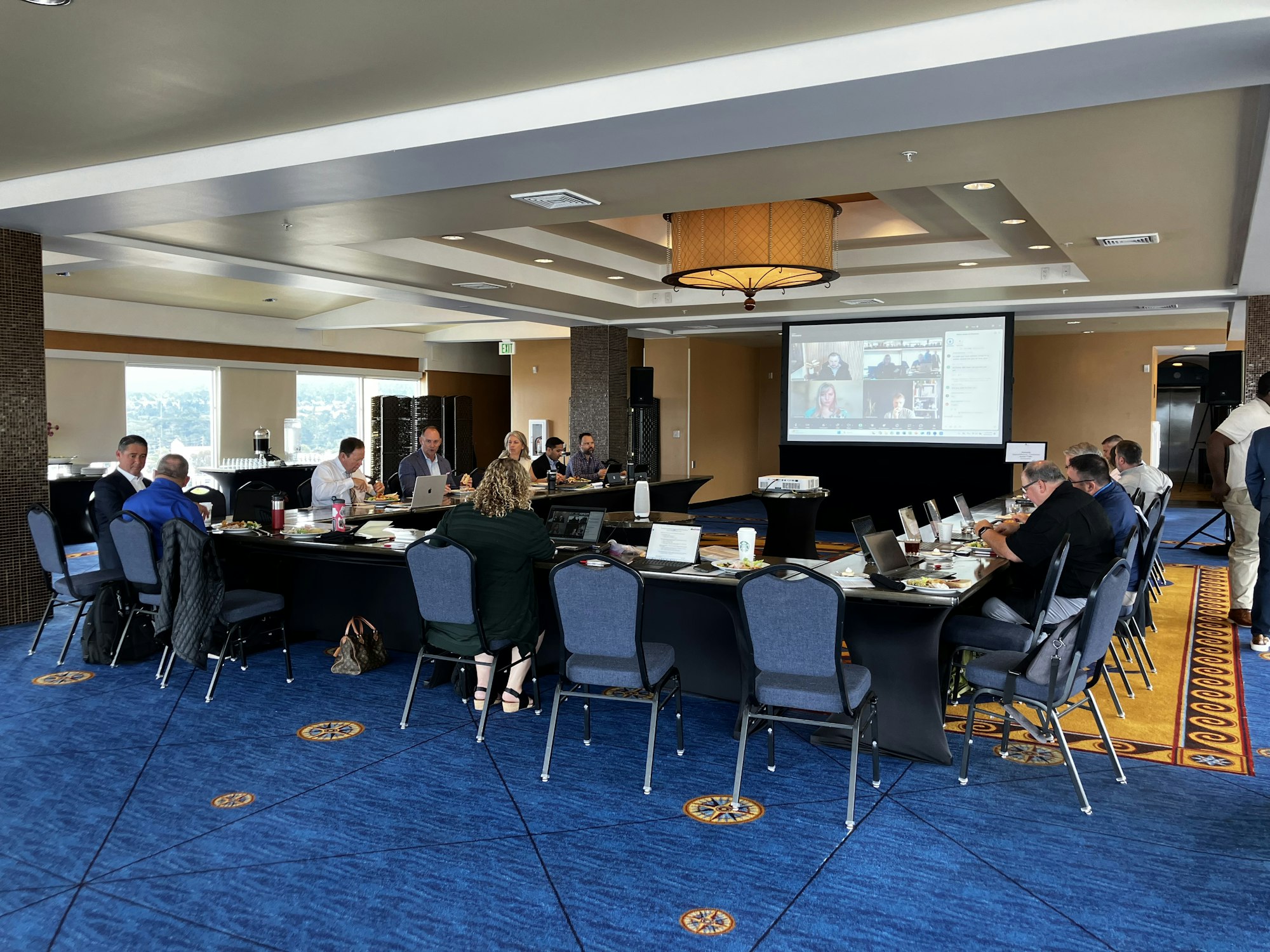 A business meeting with people seated around tables, laptops open, and a video call on a large screen.