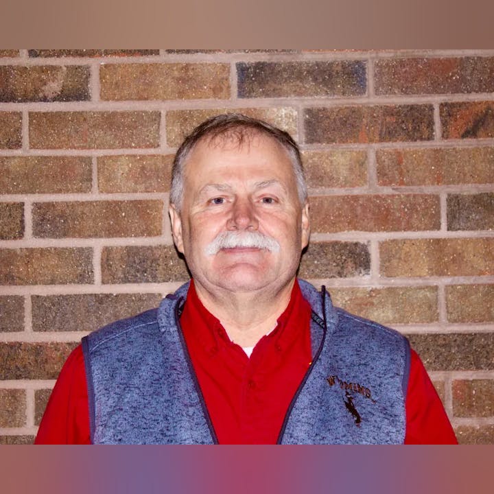 A man with a mustache smiling in front of a brick wall.