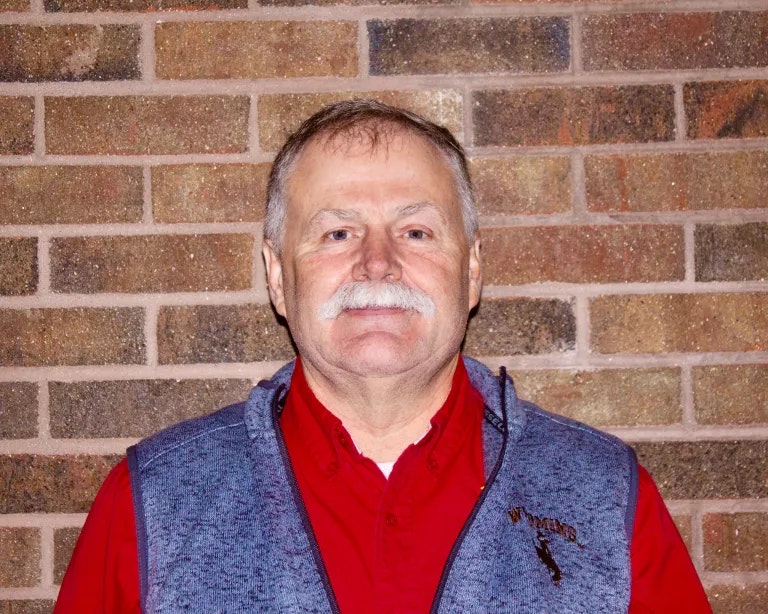 A man with a mustache smiling in front of a brick wall.