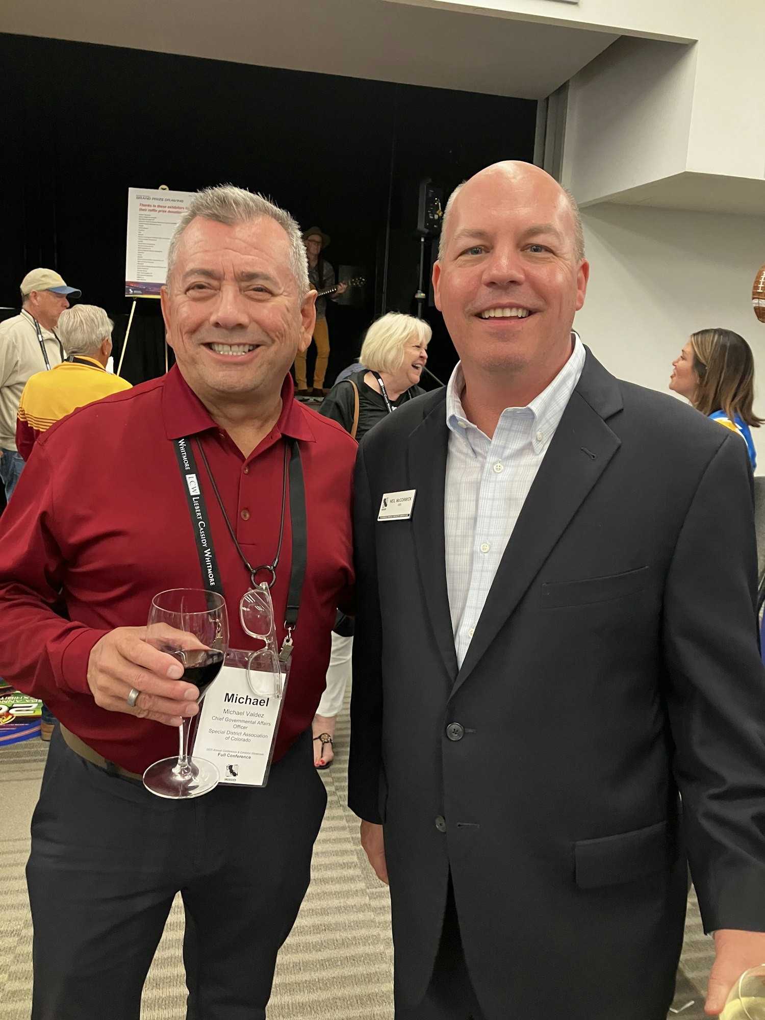 Two men smiling at a social event, one holding a wine glass, with a few people in the background.