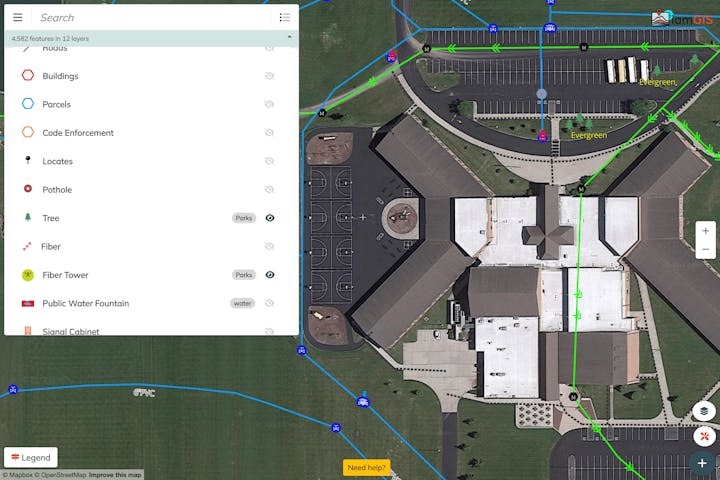 Aerial map with GIS data layers: roads, buildings, parcels, trees, fiber, and more. Shows a large building with parking and sports areas.