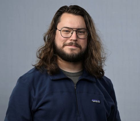 A person with long hair and glasses, wearing a blue zip-up jacket, stands against a neutral background.