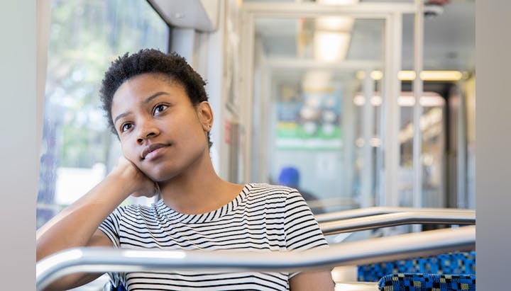 A person with short hair sits on a train, resting their head on their hand and gazing thoughtfully out of the window.