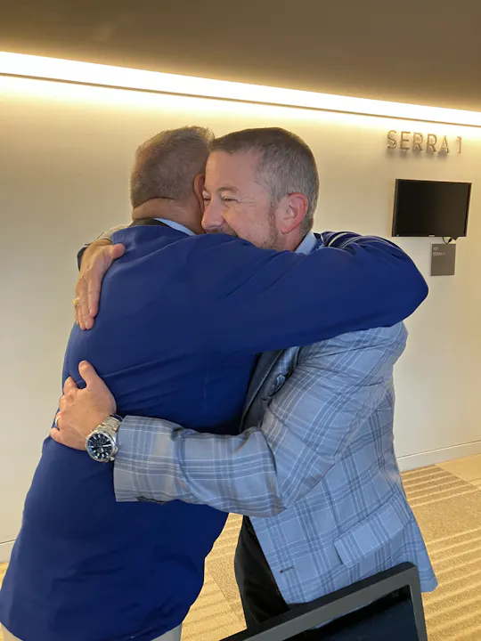 Two men are embracing in a warm, friendly hug, with a modern interior background that includes a wall sign.
