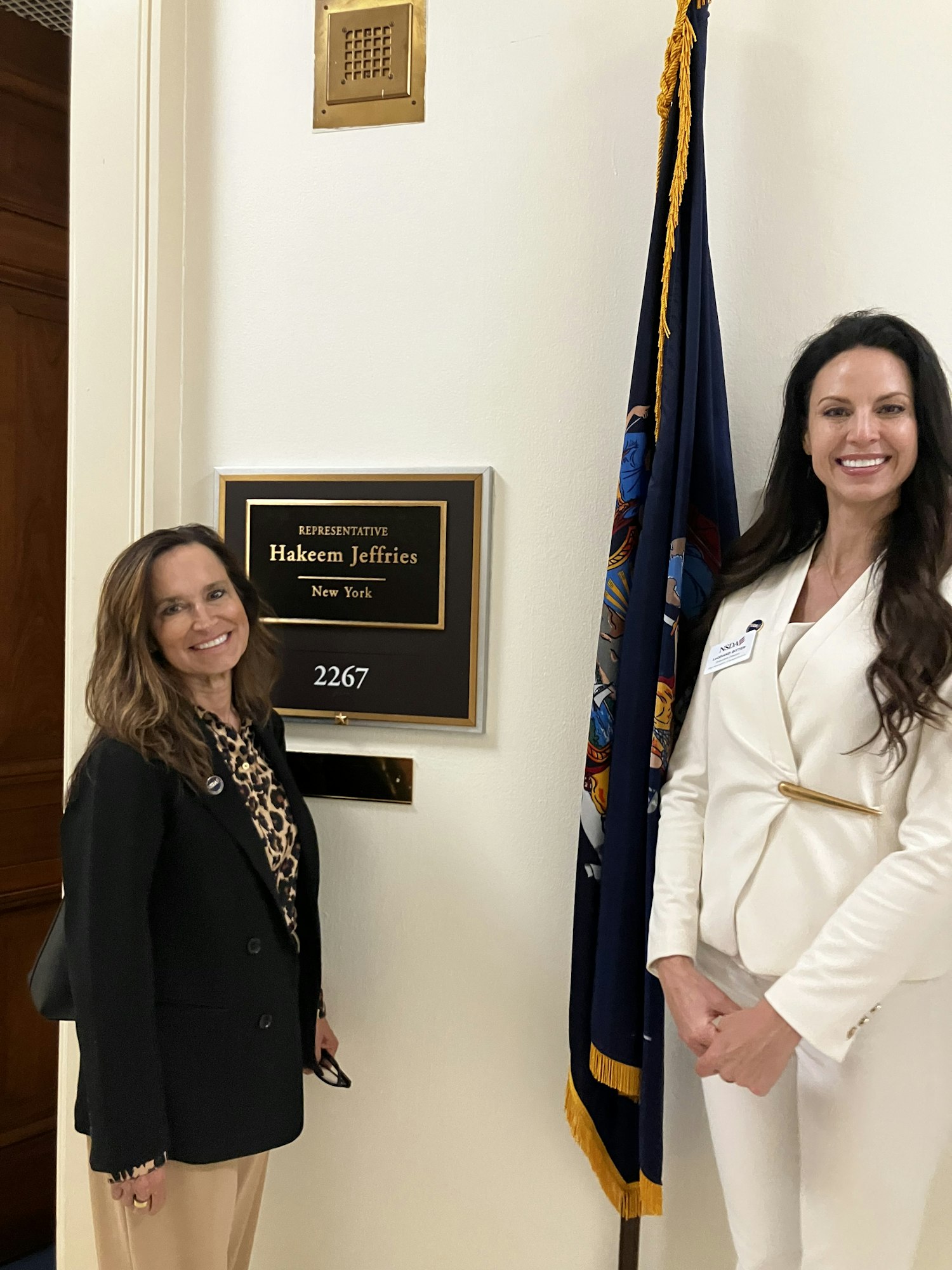Two women pose in front of a sign for Representative Hakeem Jeffries' office, with a flag in the background.