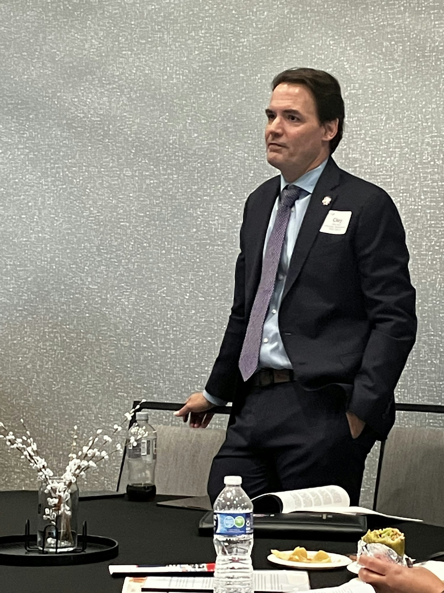 A person in formal attire stands near a table with a water bottle, food, and papers.