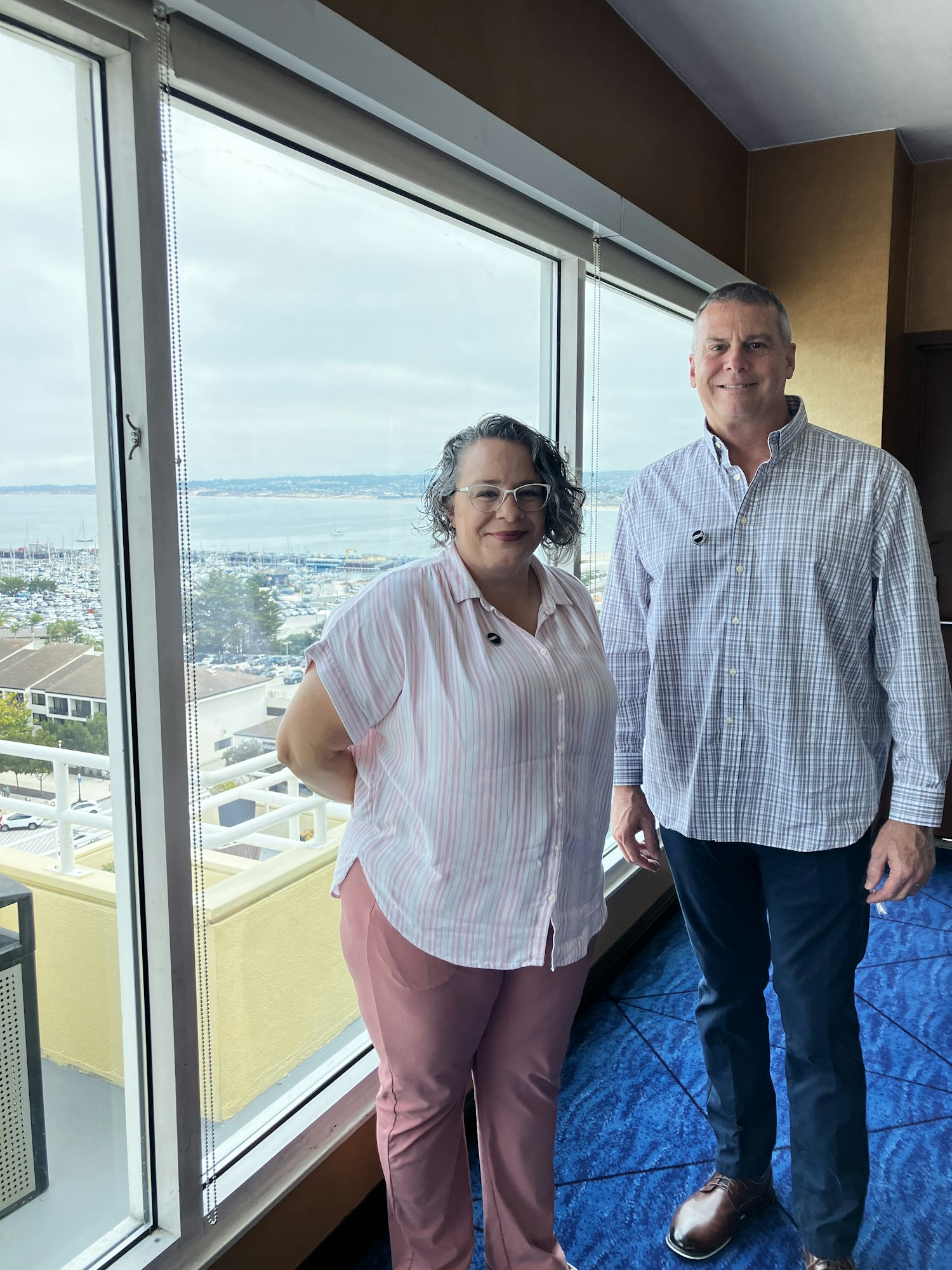 A man and woman standing indoors by a large window with a scenic view.