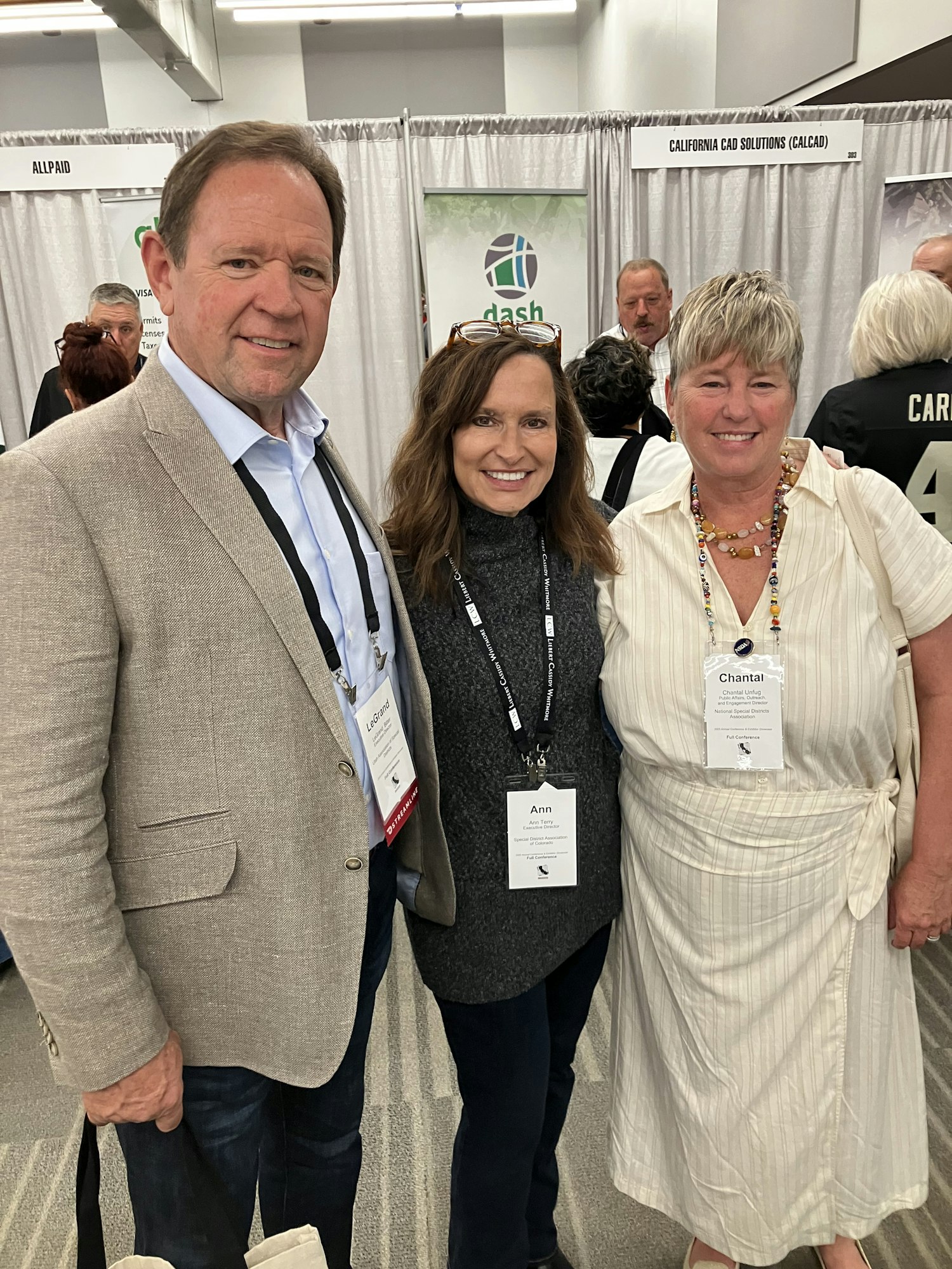 Three people smiling at a conference with name tags, standing in front of an exhibit booth.