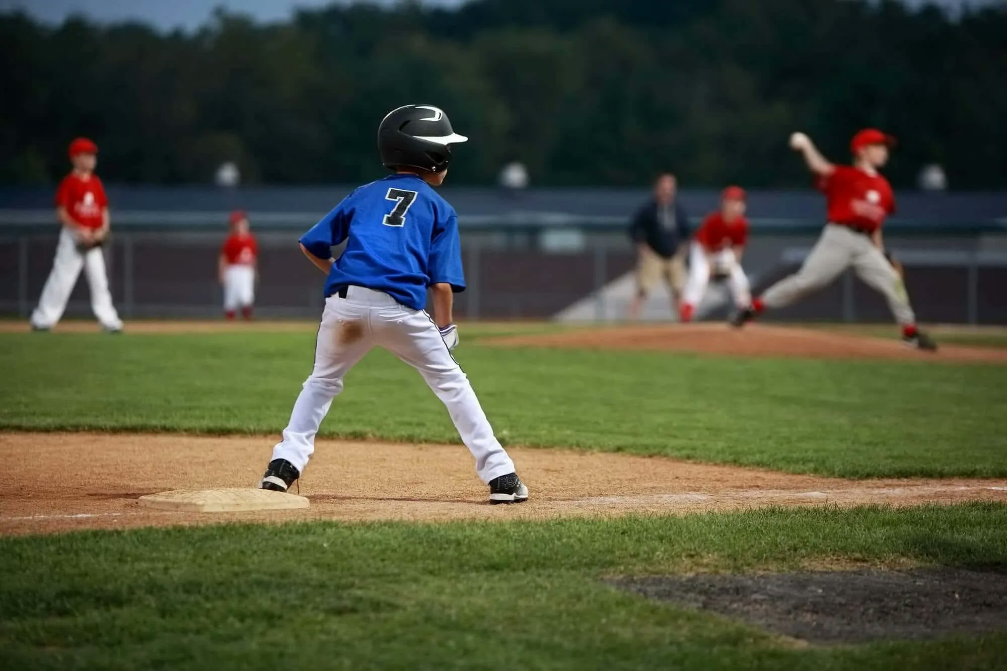 A youth baseball game, with a player in blue and others in red, focused on the pitcher mid-throw.