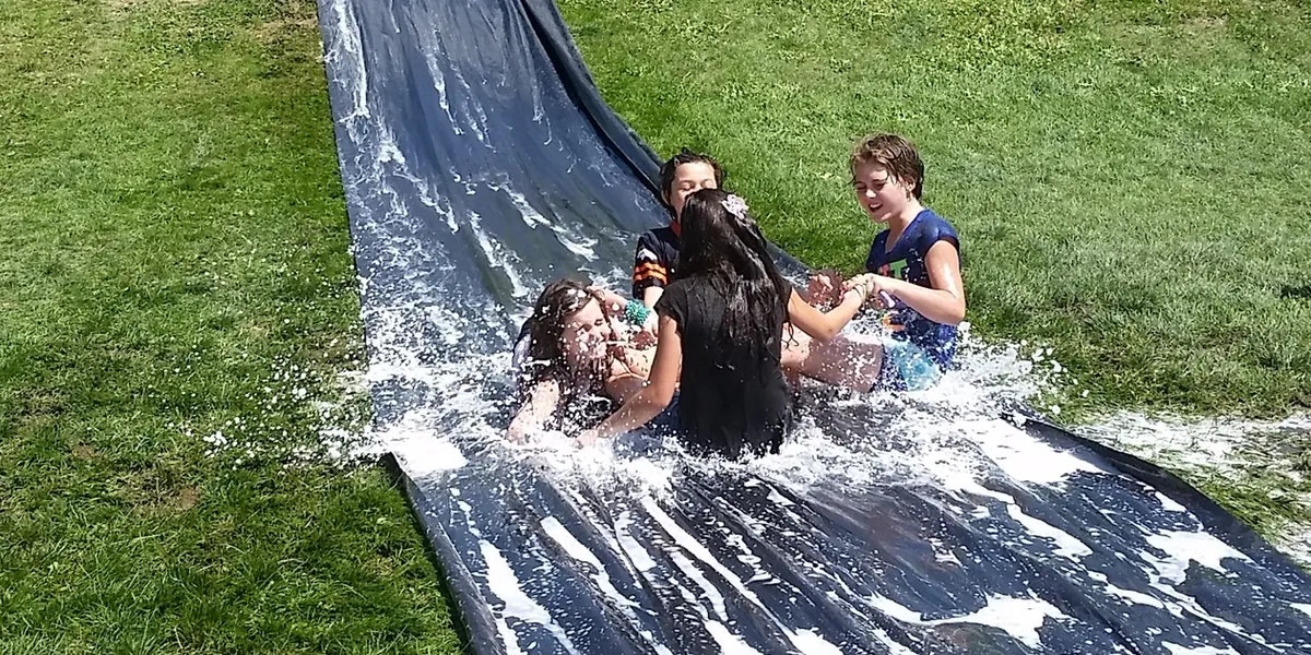 Children playing on a watery slip and slide in a grassy backyard, splashing and having fun.