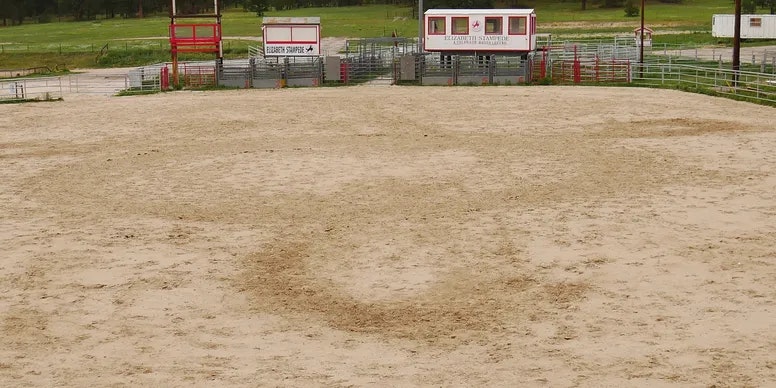 An empty rodeo arena with metal fencing, gates, and a few small buildings in the background.