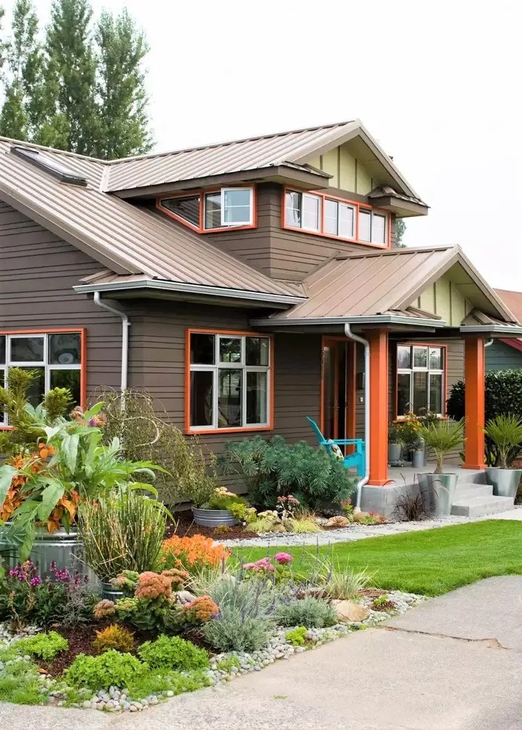 A modern house with brown siding, orange trim, and a lush front garden featuring diverse plants and greenery.