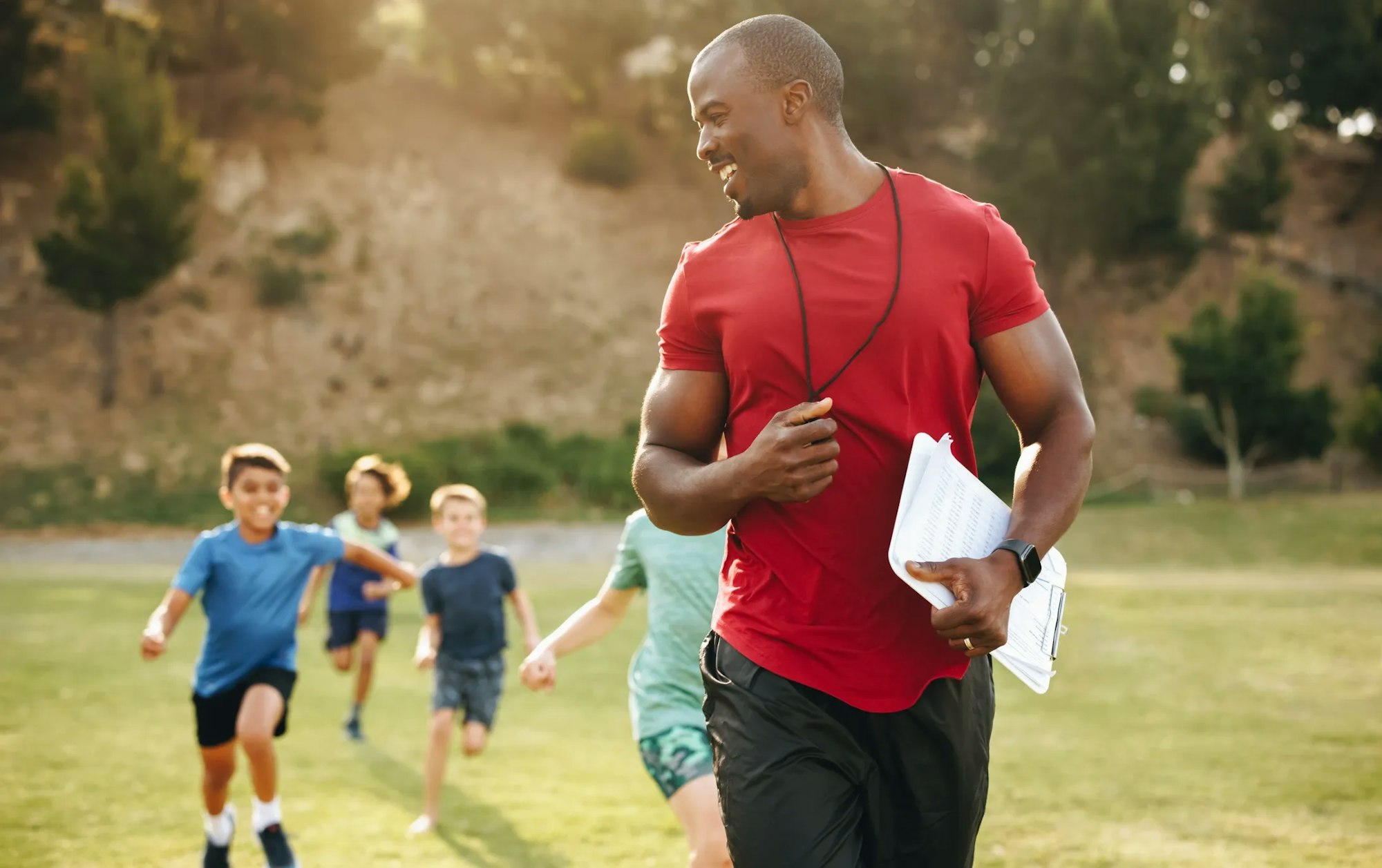 A coach with a whistle runs energetically while children follow him on a grassy field.