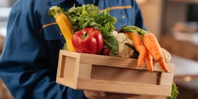 A person holding a wooden crate filled with fresh vegetables like carrots, bell peppers, and leafy greens.