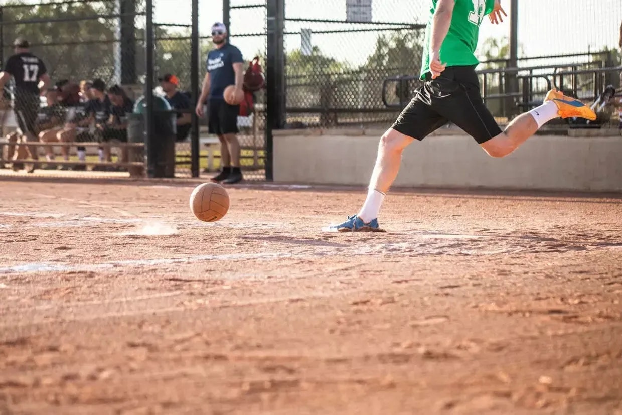A person in a green shirt kicking a ball on a dirt field, with others watching in the background.
