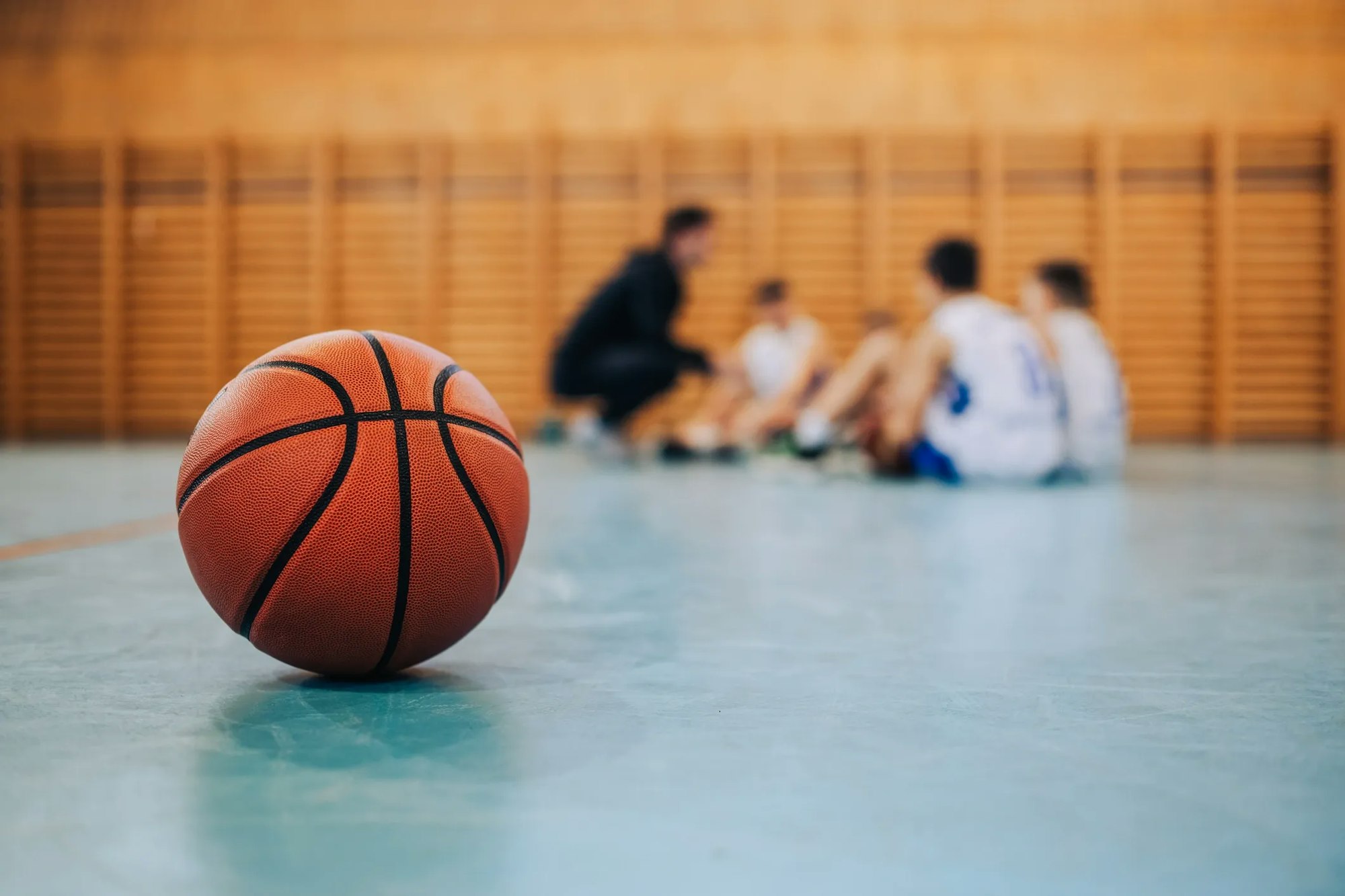 A basketball in focus on a gym floor, with blurred figures of a coach and players in the background.