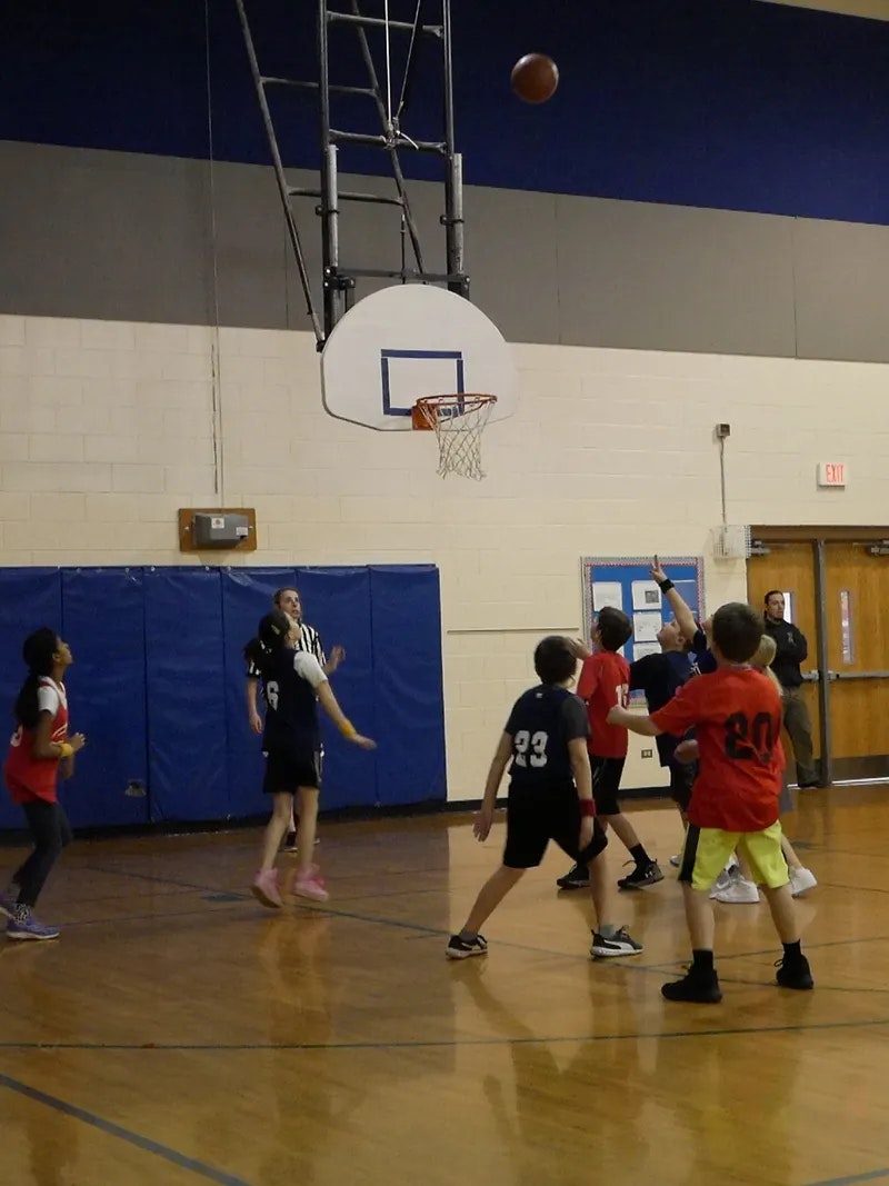 A group of kids playing basketball indoors, a ball is near the hoop.