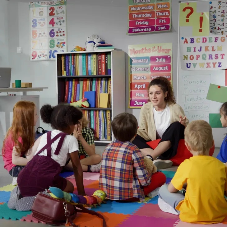 A teacher sits on the floor with young children in a classroom, surrounded by educational posters and colorful mats.