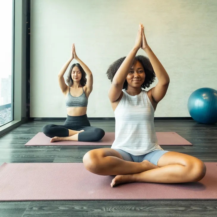 Two people practicing yoga in a studio, seated with hands in prayer position, exercise mat and ball visible.