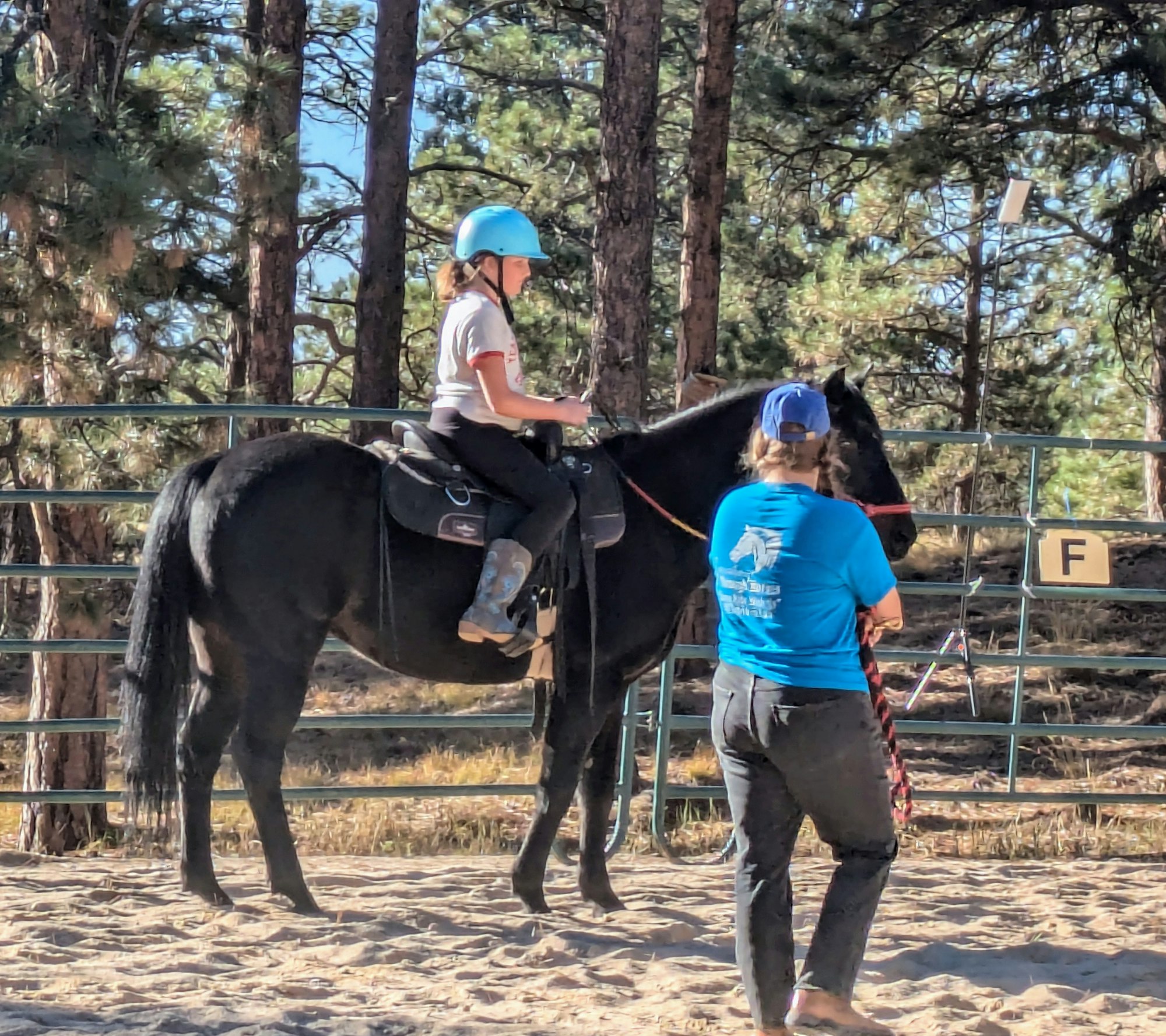 A girl in a blue helmet is riding a black horse while an instructor guides her in a sandy, wooded arena.