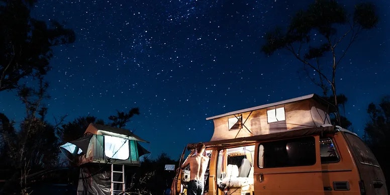 Starry night sky above a camper van with a pop-up roof and a person standing outside; tent set up nearby among trees.