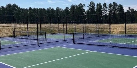 Outdoor pickleball courts with nets and fencing, surrounded by trees and open field.