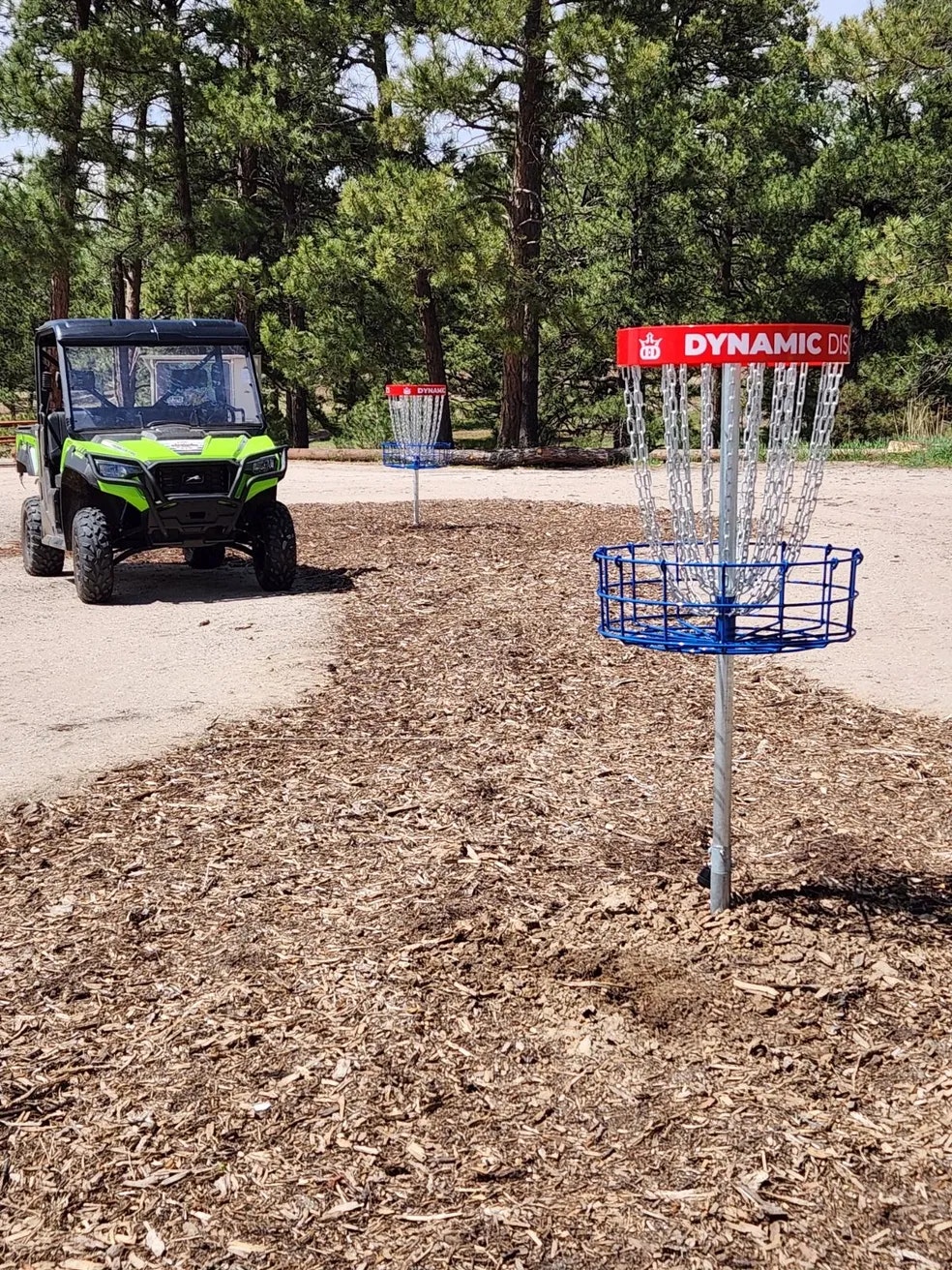 Disc golf baskets on a course with wood chip ground, a green utility vehicle nearby, and trees in the background.