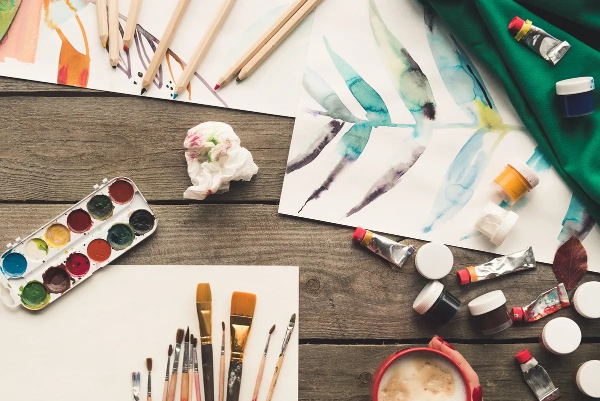 Art supplies scattered on a wooden table: paintbrushes, watercolors, papers with paintings, pencils, paint tubes, and a cup of coffee.
