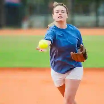 Person playing softball, wearing a blue shirt, holding a softball and glove, on an orange dirt field.