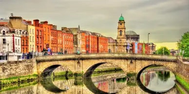 A stone bridge spans a river, with colorful buildings and a clock tower in the background under a cloudy sky.