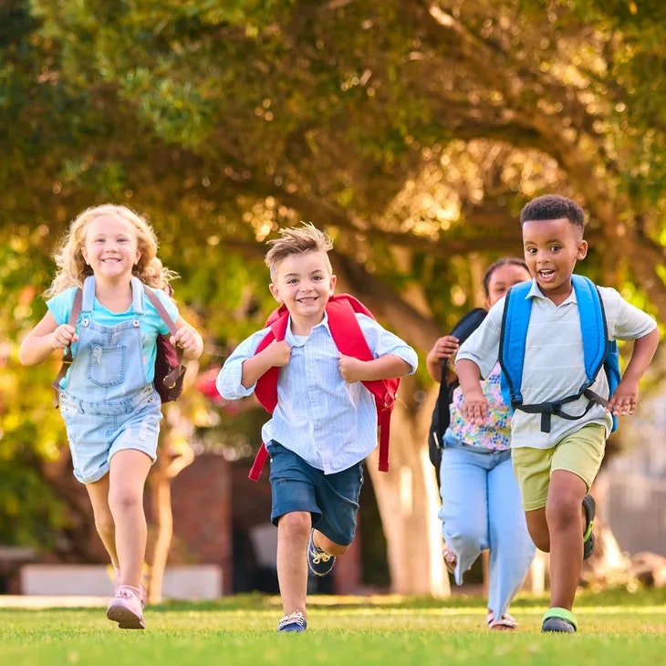 Four children with backpacks running happily on grass, surrounded by trees.