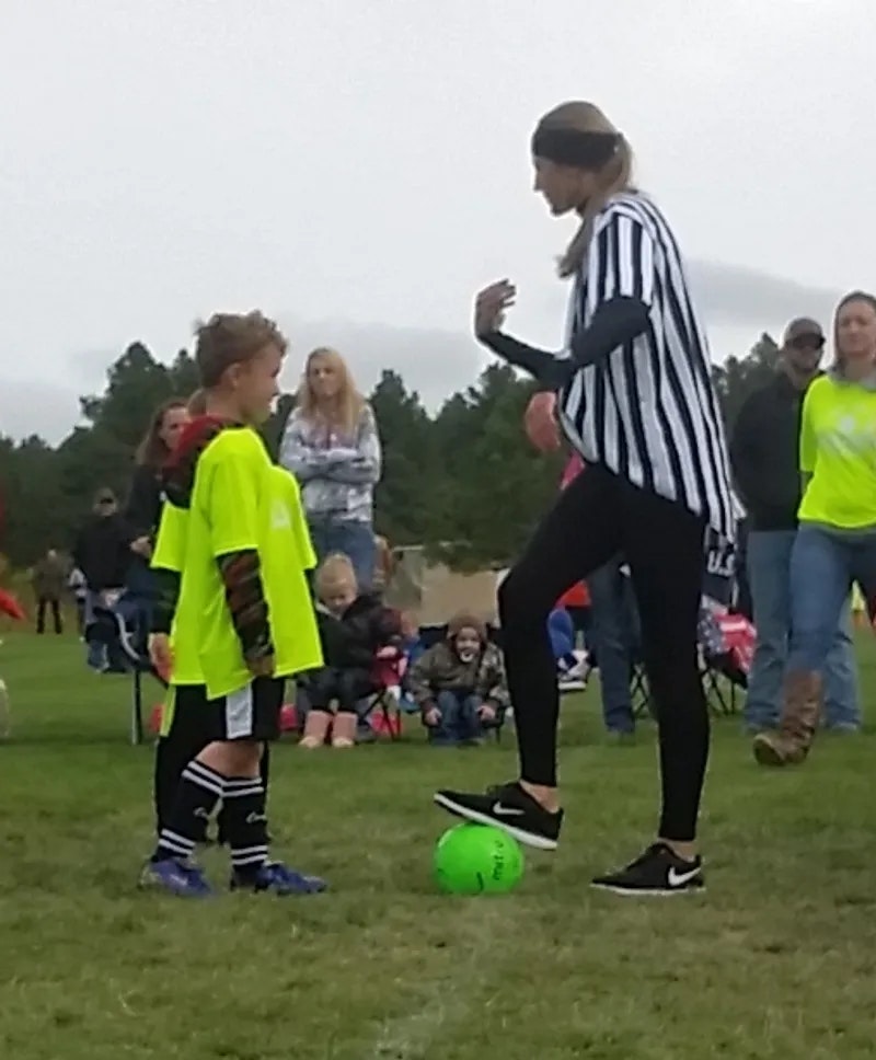 A referee is talking to a young soccer player on a field, with a green ball on the ground and spectators in the background.