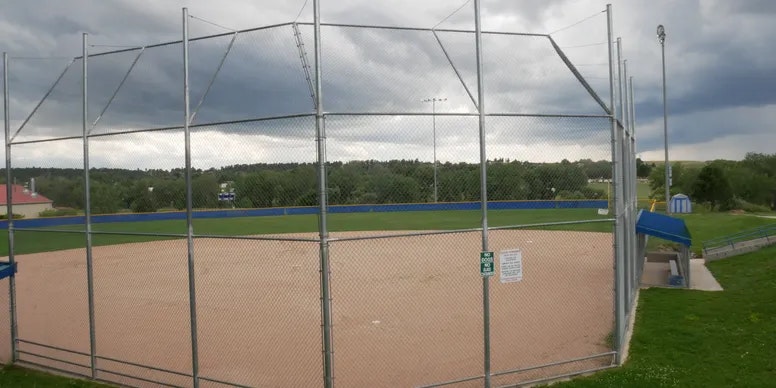 A baseball field with a fenced backstop, empty spectator stands, and a cloudy sky.