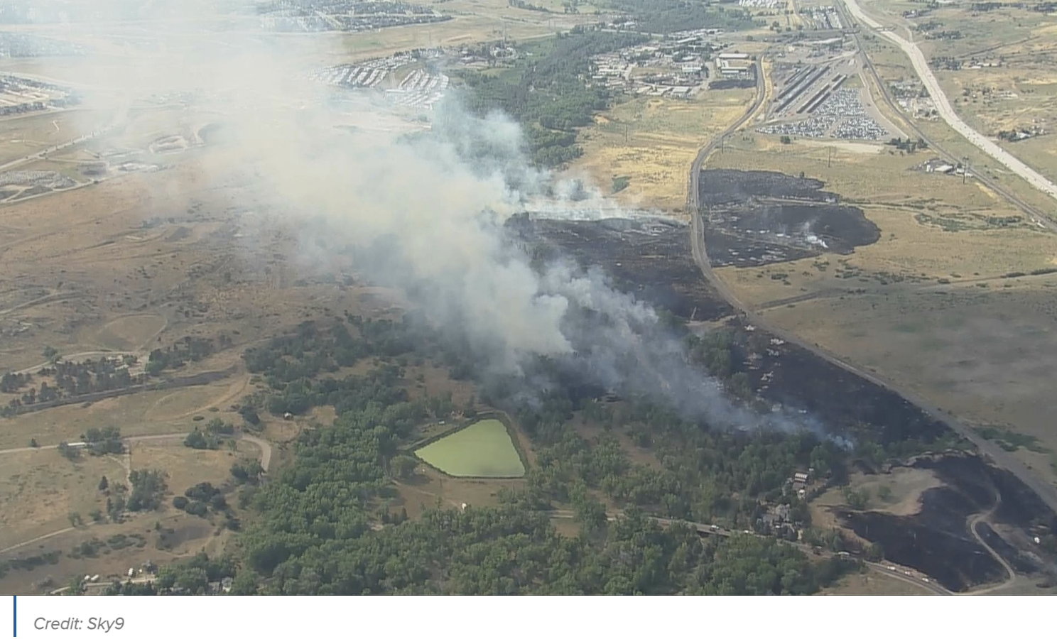Aerial view of a wildfire with smoke billowing over a landscape, partially burned fields, and nearby roads.