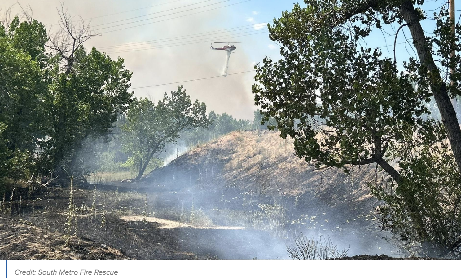 Helicopter dropping water over a smoldering field, trees nearby, and smoke rising.