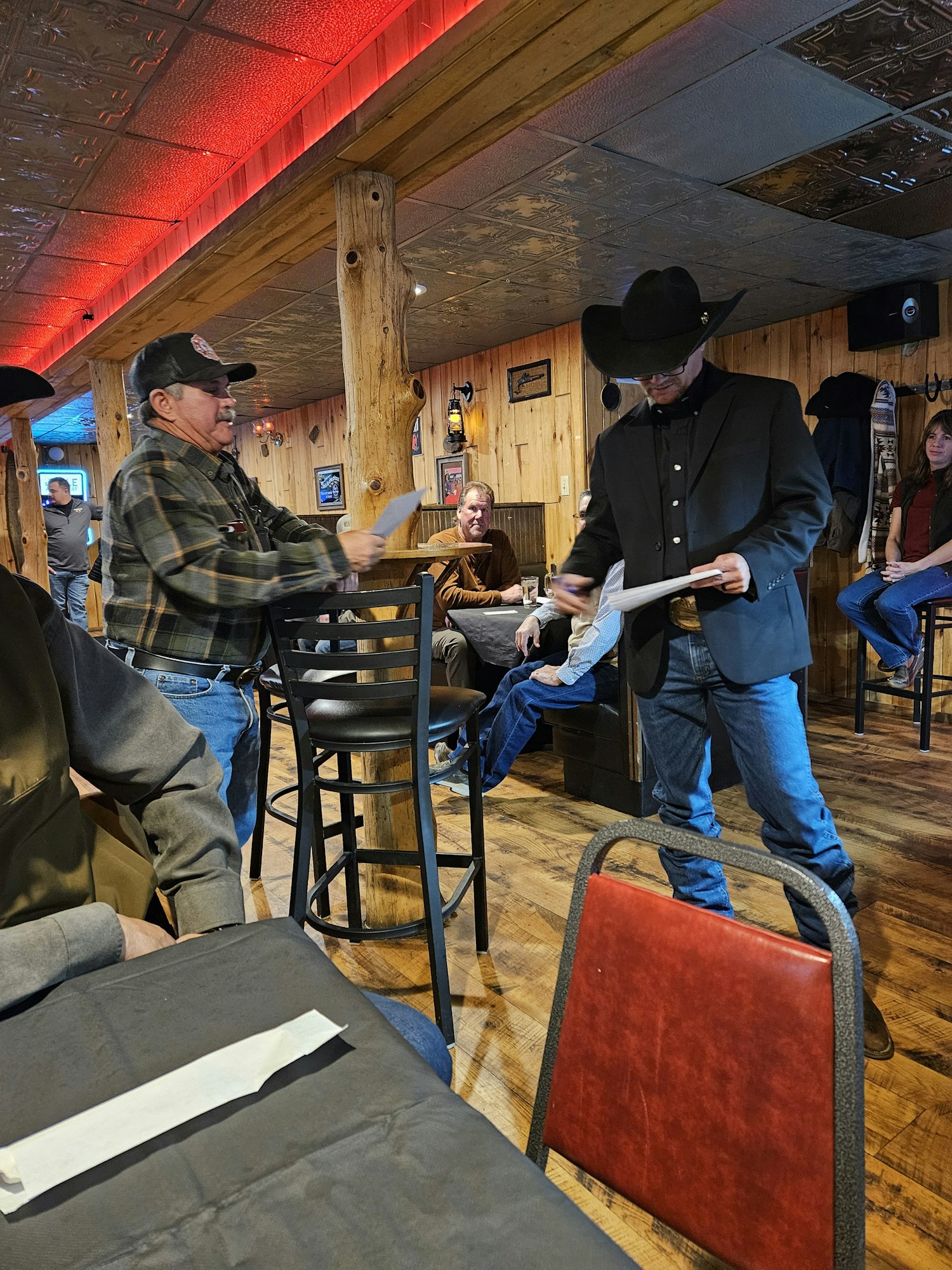 A rustic interior with people, wooden walls, a man in a cowboy hat, and dining furniture. (Note: Image is rotated)