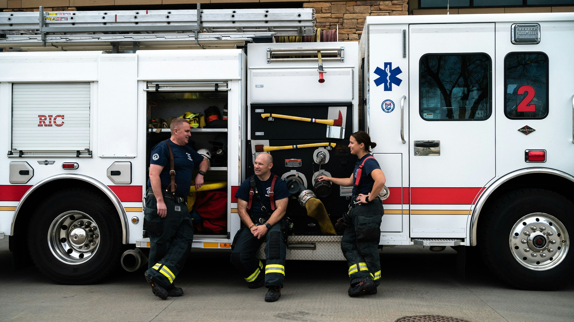 Three firefighters with a fire truck, two seated and chatting, one standing, gears in background.