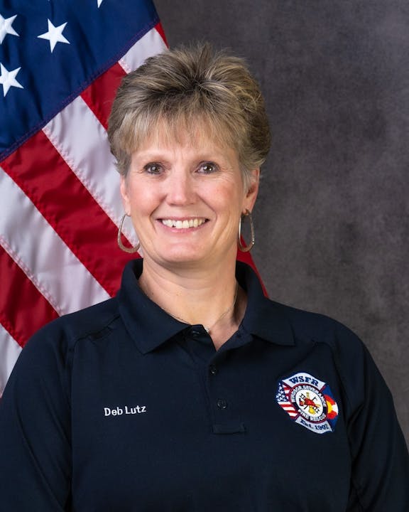 Person with badge, "Deb Lutz" name tag, American flag backdrop.