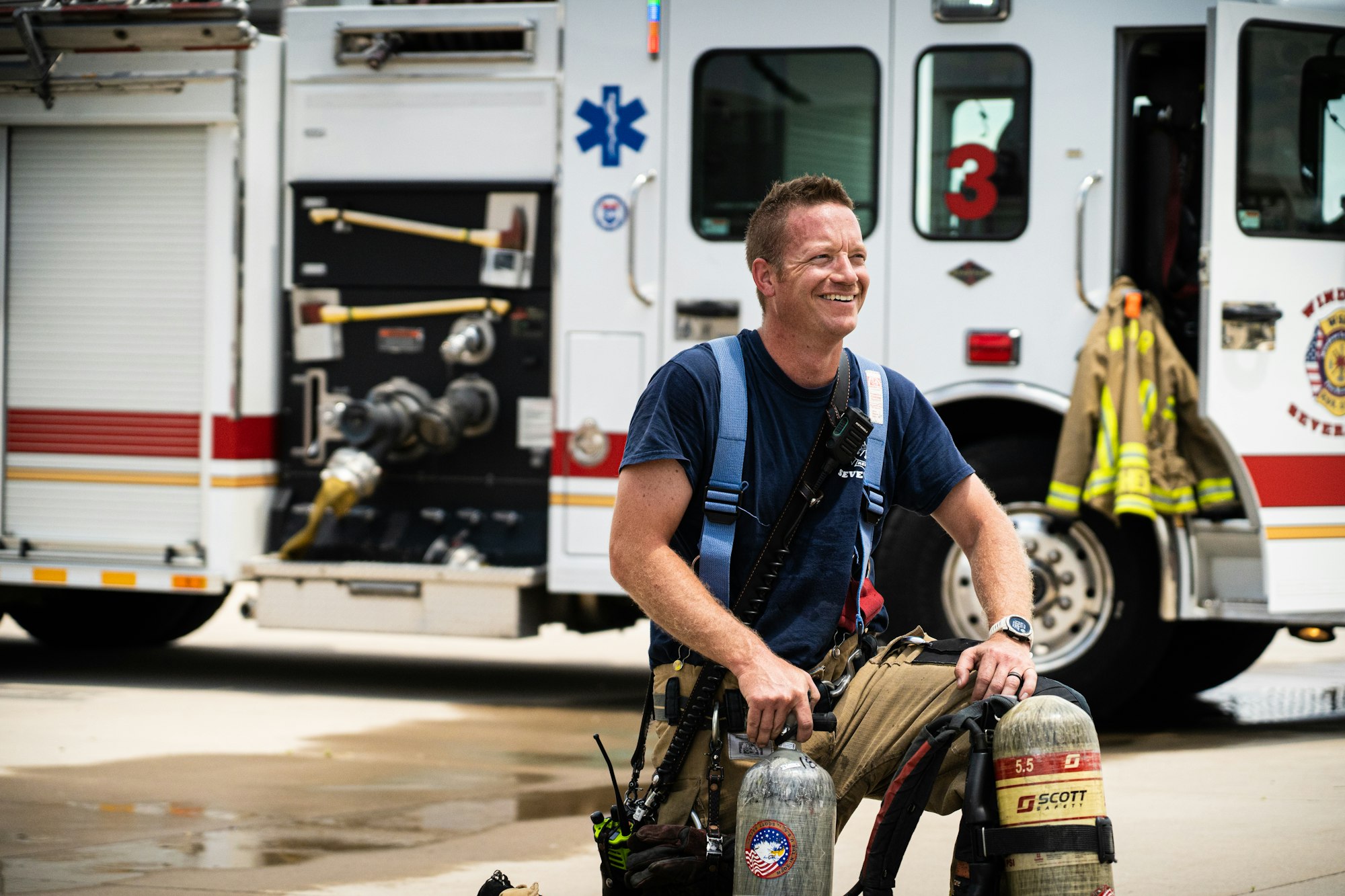 A smiling firefighter with gear in front of a fire truck.