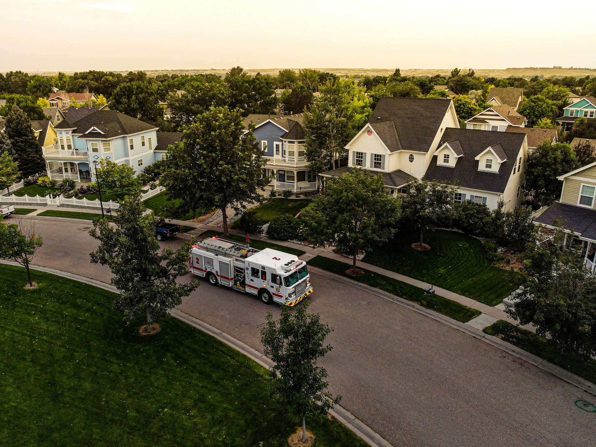 An aerial view of a suburban neighborhood with a fire truck on the street and houses surrounded by trees.