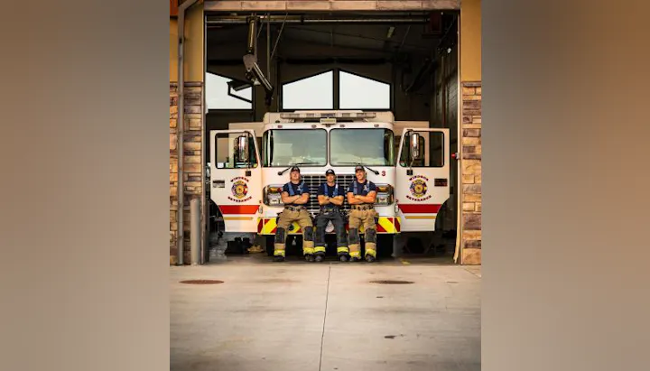 Three firefighters sitting in front of a fire truck in a fire station bay.