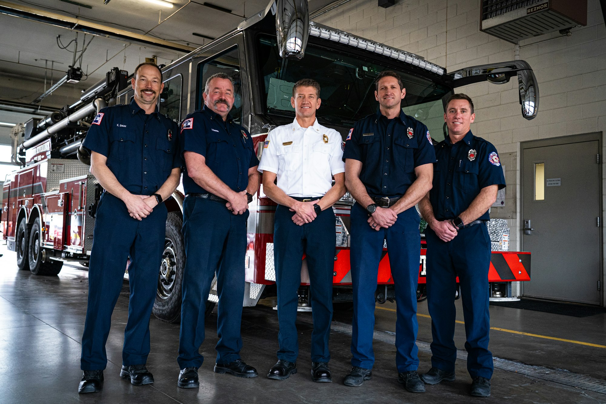 Five firefighters in uniform standing in front of a firetruck inside a fire station.