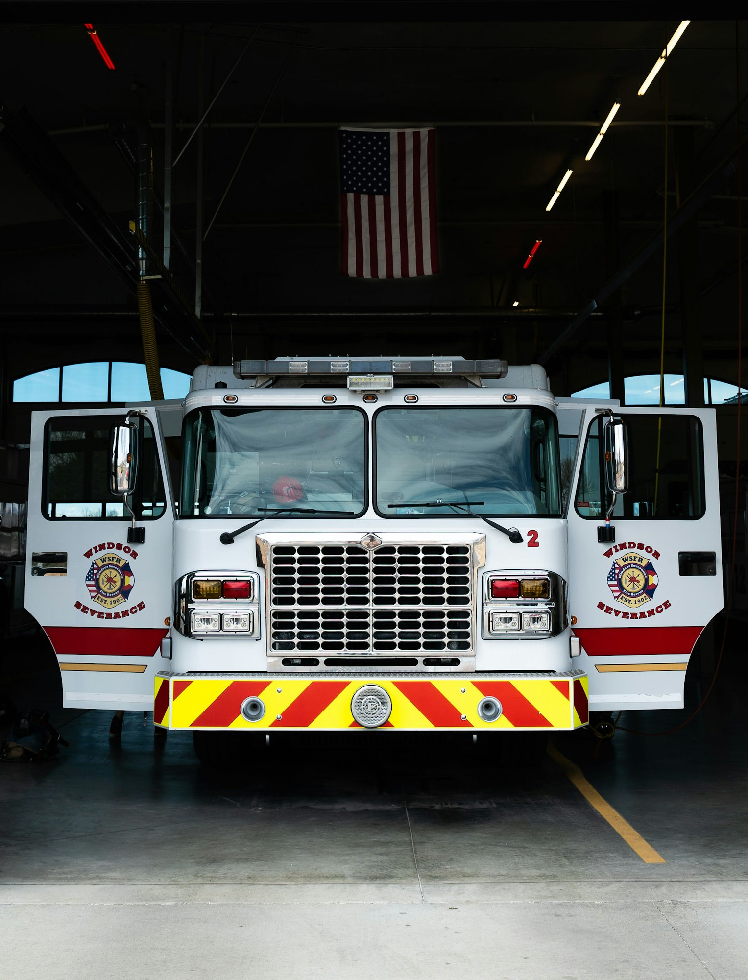 A fire truck parked inside a station, with an American flag hanging in the background.