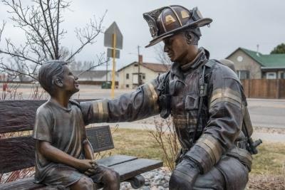 A firefighter in gear touching the hand of a bronze statue of a child on a bench, creating a poignant scene.