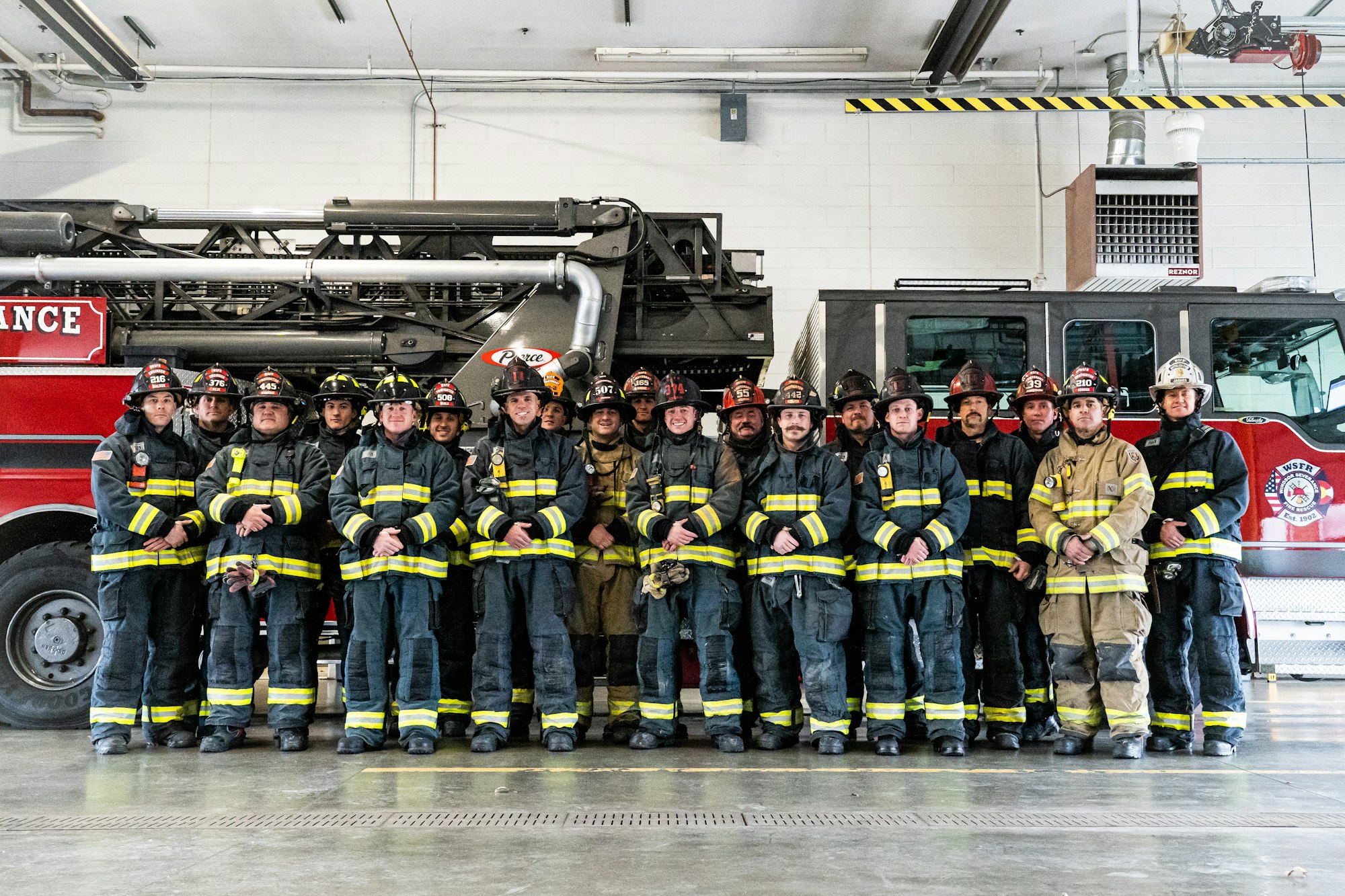 A group of firefighters in gear posing in front of a fire truck inside a fire station.